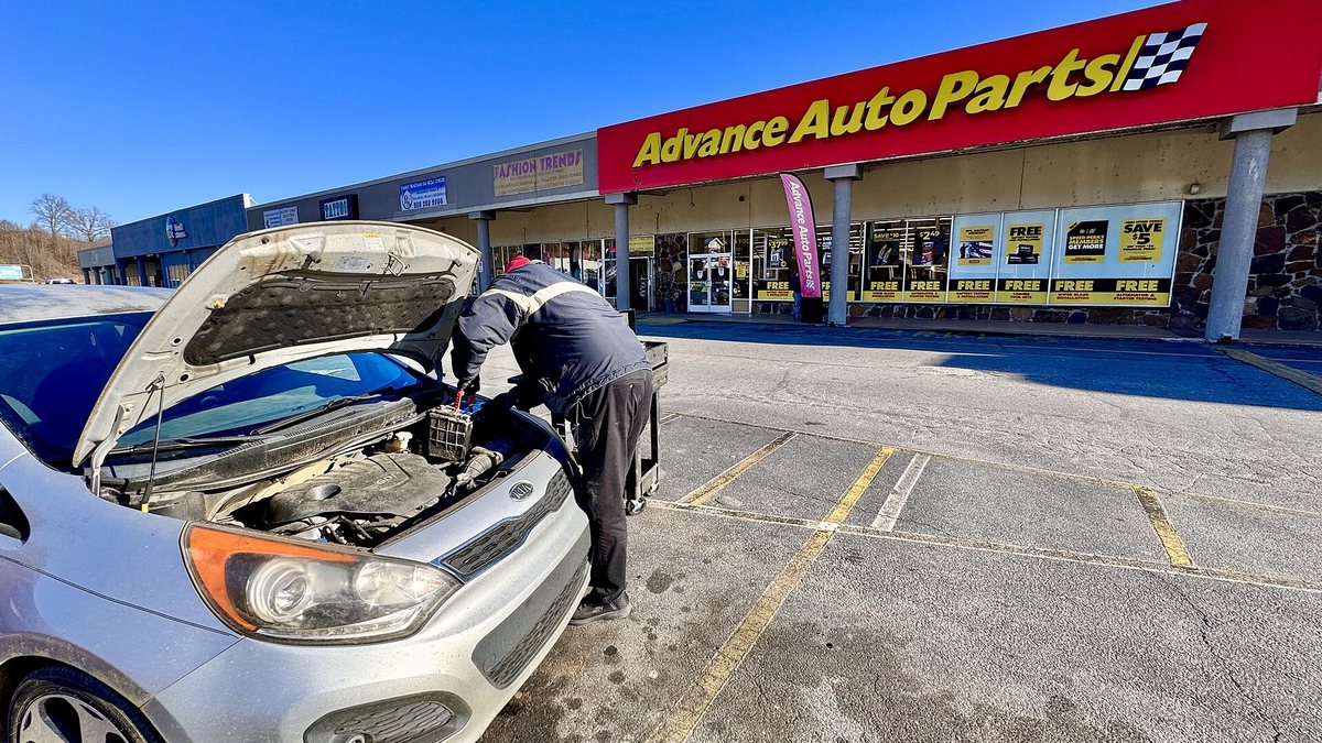 An Advance Auto Parts store employee changes a car battery in a parking lot in front of the shop